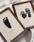 Two framed prints with black hand and footprints on beige fabric backgrounds.