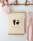 Decorative wall hanging with hand and footprints on a beige background, hanging on a gold hook with pink ribbon.